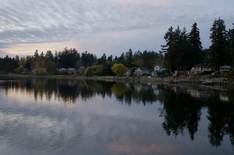 A still lake reflecting a wooded shoreline and houses under a cloudy sky