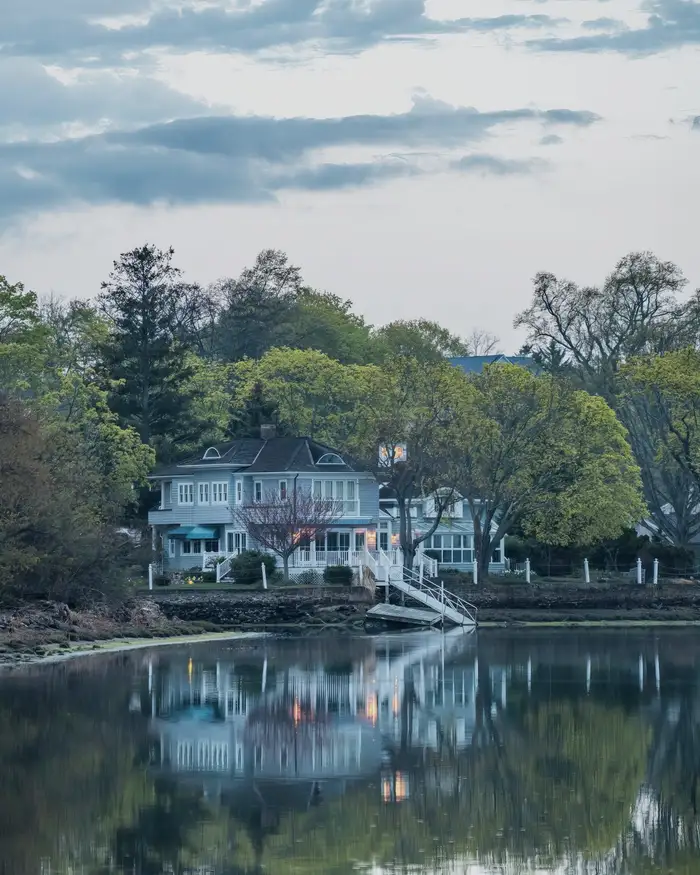 A white lake house among trees reflected in still blue water