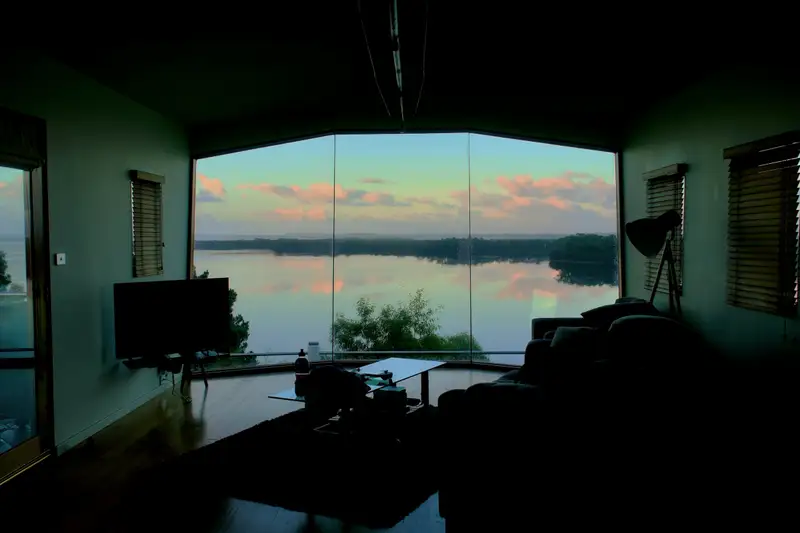 A dark room with a wide window looking out over a lake at dusk
