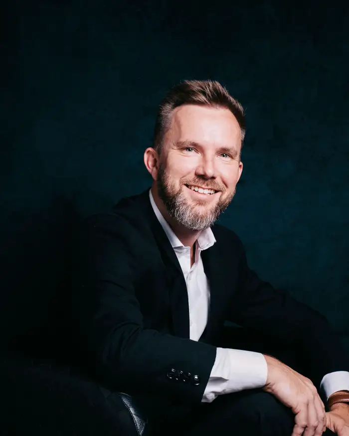 Sample portrait: a man in a dark suit seated, smiling, against a dark teal backdrop